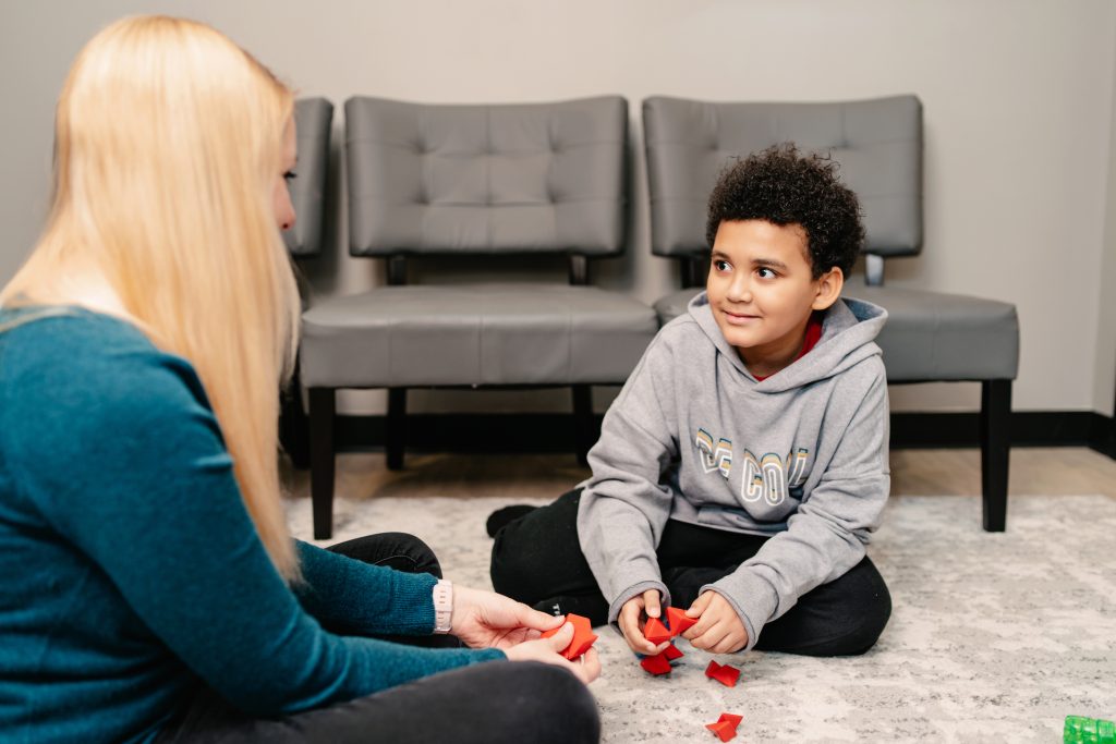 A young boy in a gray hoodie smiles while playing with red puzzle pieces on the floor with a female ABA therapist.